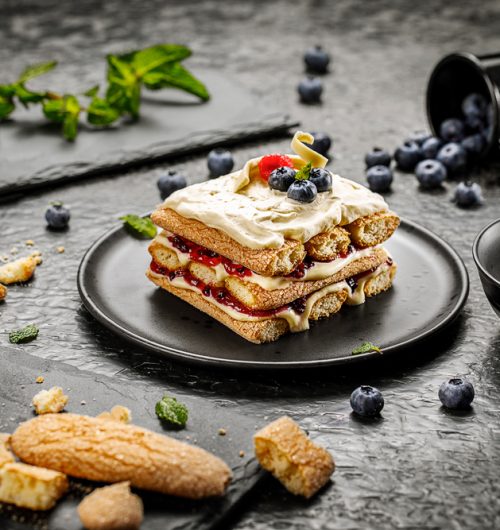 Biscoitos champanhe dourados empilhados em prato preto, cobertos com nata e frutas vermelhas, sobre mesa escura.