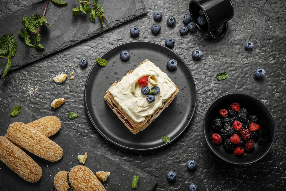 Biscoitos champanhe dourados empilhados em prato preto, cobertos com nata e frutas vermelhas, sobre mesa escura.