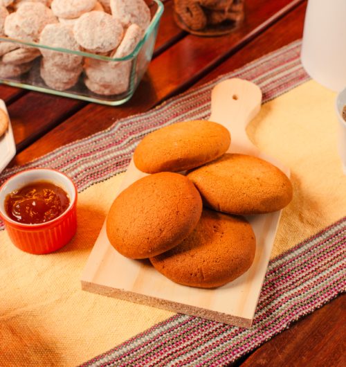 Biscoito bolachão grande e redondo, de cor dourada e textura crocante, colocado sobre mesa de madeira ao lado de xícara de café.
