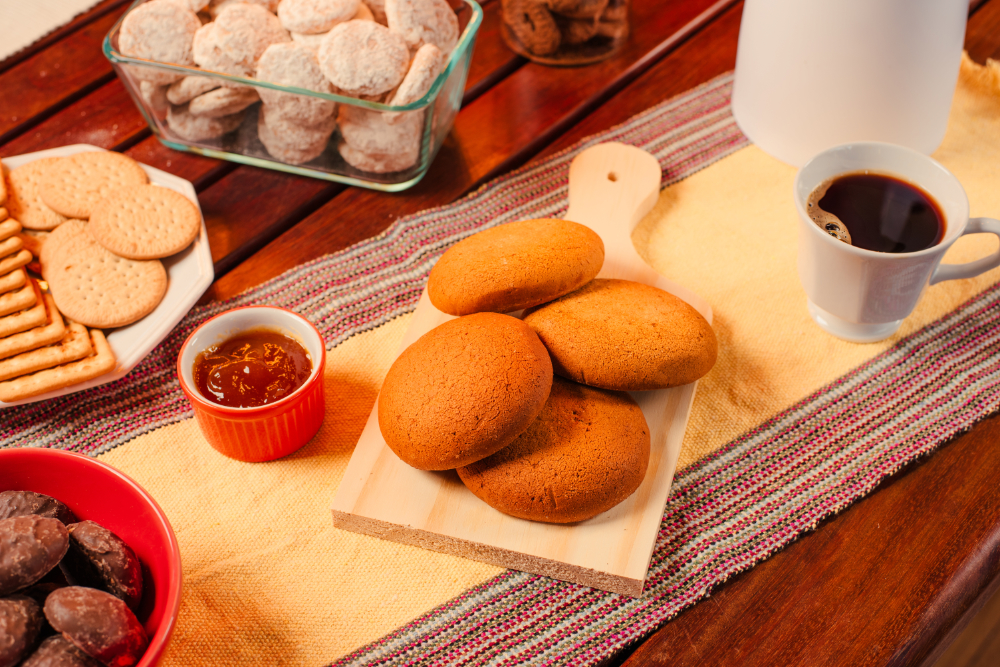 Biscoito bolachão grande e redondo, de cor dourada e textura crocante, colocado sobre mesa de madeira ao lado de xícara de café.