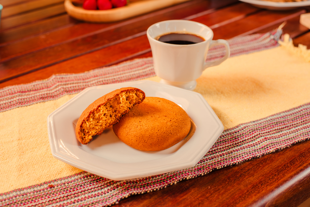 Biscoito bolachão grande e redondo, de cor dourada e textura crocante, colocado sobre mesa de madeira ao lado de xícara de café.