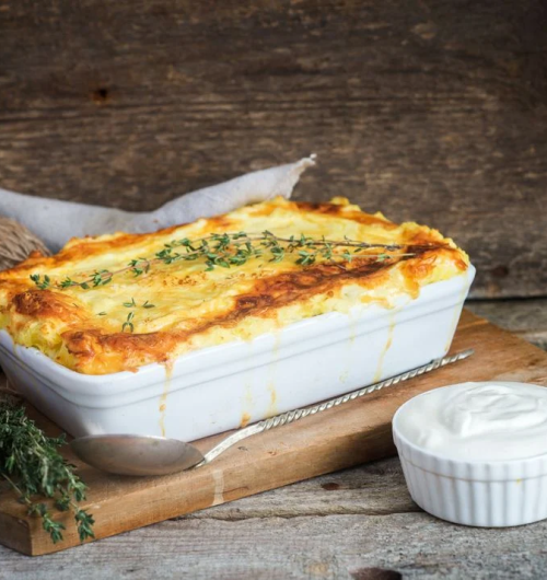 Travessa branca com batatas gratinadas, cobertas por queijo derretido, servida sobre mesa de madeira.