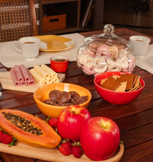 Mesa de café da manhã com xícara de chá, pratos, queijo, presunto, rosquinhas e biscoitos, acompanhados de frutas.