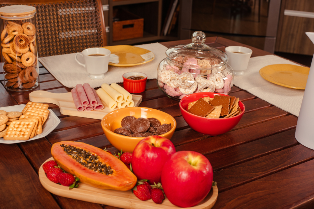 Mesa de café da manhã com xícara de chá, pratos, queijo, presunto, rosquinhas e biscoitos, acompanhados de frutas.