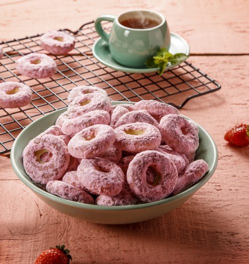 Pote com biscoito calda sabor morango, exibido sobre fundo rosa e xícara de chá