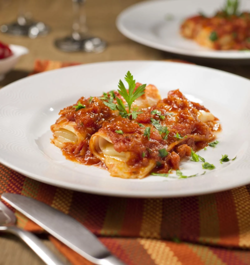 Prato branco com canelones recheados, servidos com molho cremoso e ervas, sobre mesa de madeira.