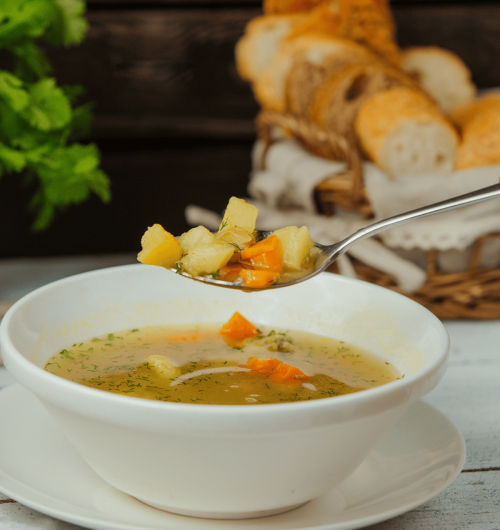Tigela branca com canja de galinha contendo cenoura, macarrão e caldo, servida sobre mesa de madeira.