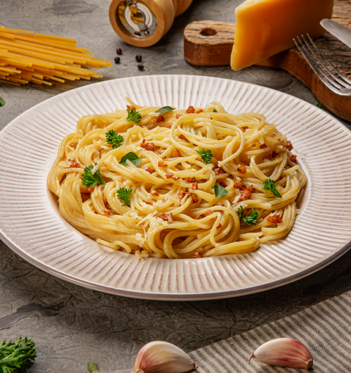 Espaguete cozido servido em prato branco, coberto com queijo ralado, bacon e manjericão fresco, sobre fundo claro e limpo.