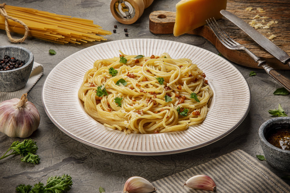 Espaguete cozido servido em prato branco, coberto com queijo ralado, bacon e manjericão fresco, sobre fundo claro e limpo.