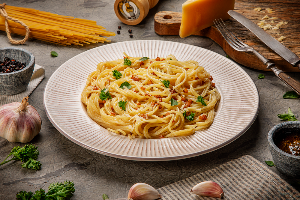 Espaguete cozido servido em prato branco, coberto com queijo ralado, bacon e manjericão fresco, sobre fundo claro e limpo.