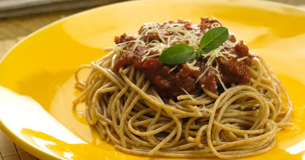 Prato com espaguete integral coberto por molho bolonhesa, queijo ralado e manjericão, servido em prato amarelo.