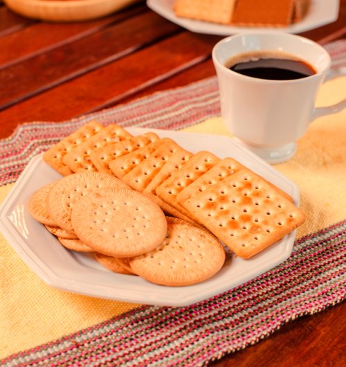 Biscoitos Maria e cream cracker, servidos em prato branco, sobre a mesa de madeira com xícara de café
