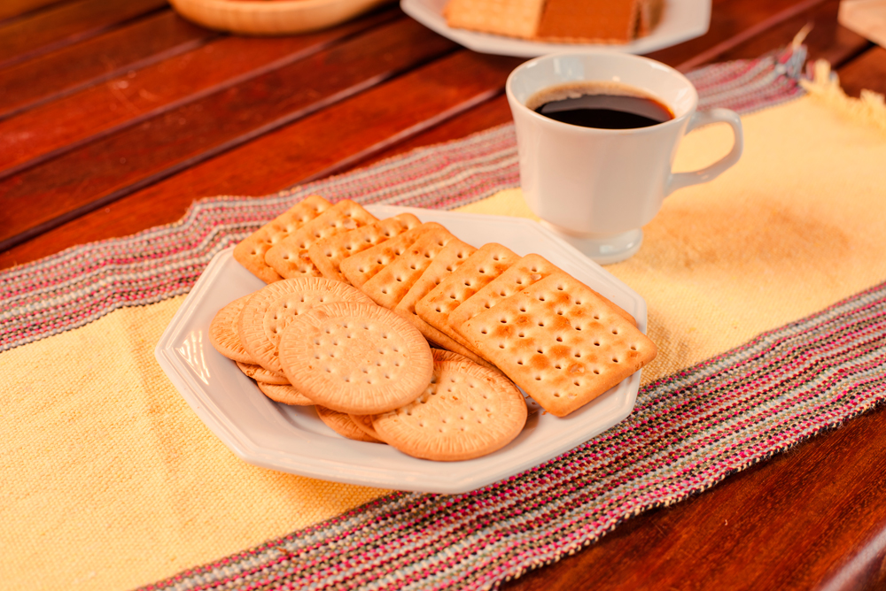 Biscoitos Maria e cream cracker, servidos em prato branco, sobre a mesa de madeira com xícara de café