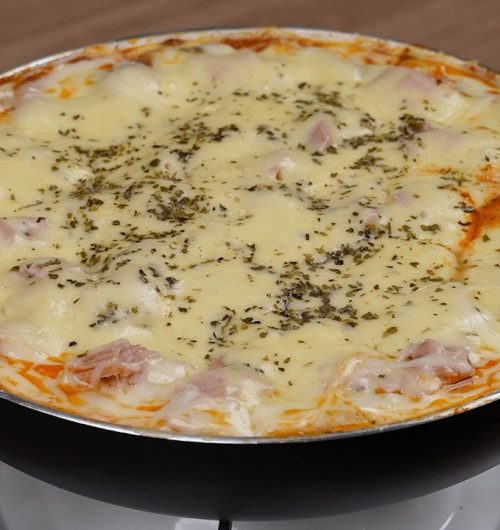 Frigideira com lasanha coberta em molho vermelho, presunto e queijo derretido, servida sobre mesa de madeira.