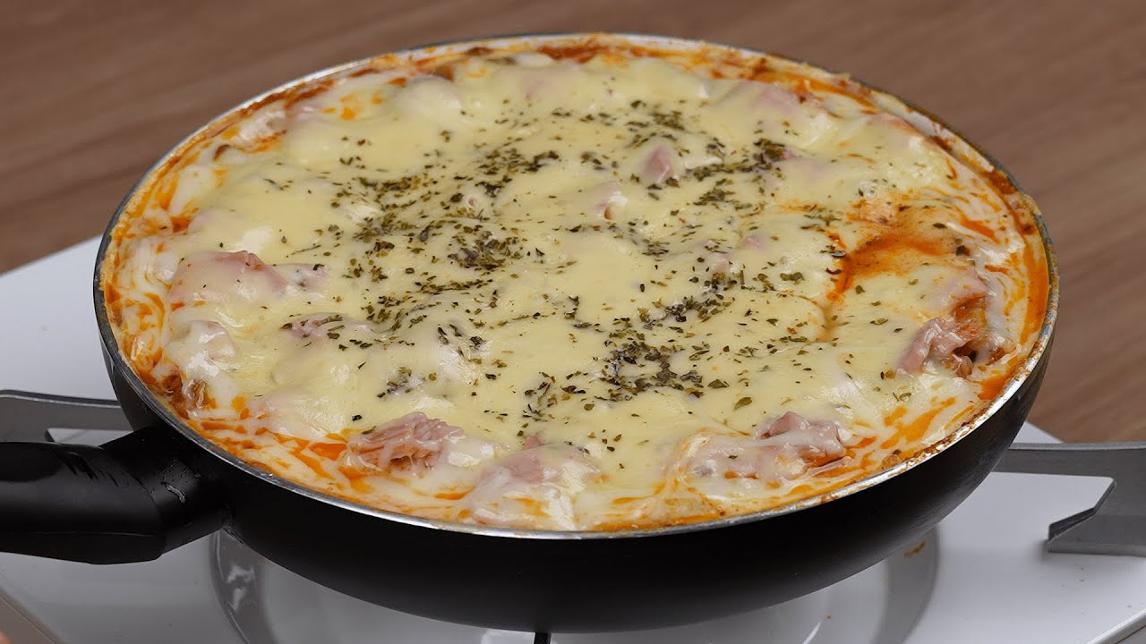 Frigideira com lasanha coberta em molho vermelho, presunto e queijo derretido, servida sobre mesa de madeira.