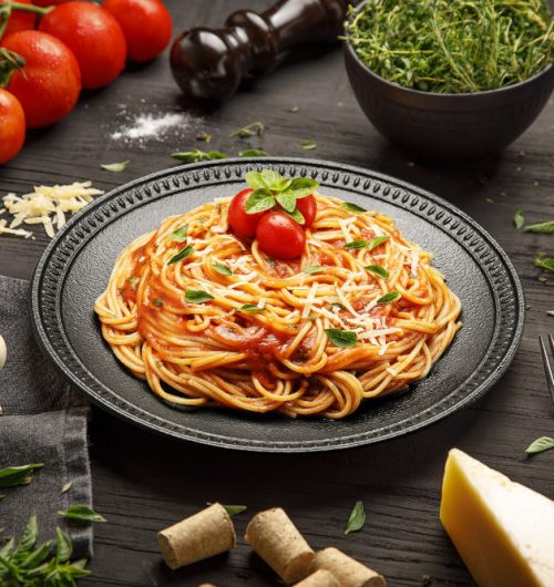 Prato de espaguete coberto com molho vermelho, tomate, manjericão e queijo ralado, em um prato escuro sobre fundo de madeira.