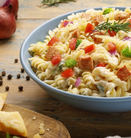 Prato fundo com macarrão parafuso com frango, cebola roxa, tomates, brócolis e queijo, sobre mesa de clara.