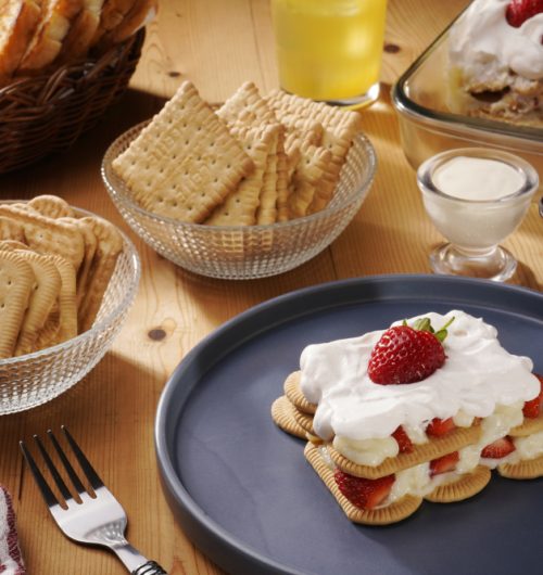 Mesa composta por biscoitos maisena, acompanhados de copo de leite, geleia, pãos tostados e sobremesa de morango, dispostos sobre mesa de madeira.