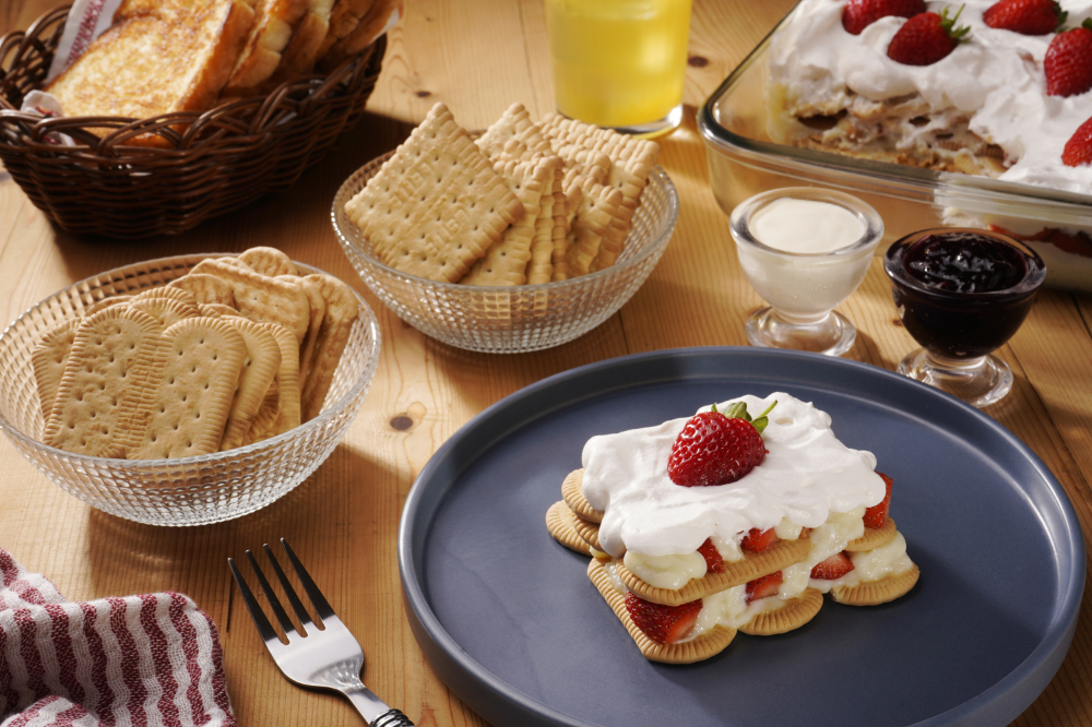 Mesa composta por biscoitos maisena, acompanhados de copo de leite, geleia, pãos tostados e sobremesa de morango, dispostos sobre mesa de madeira.