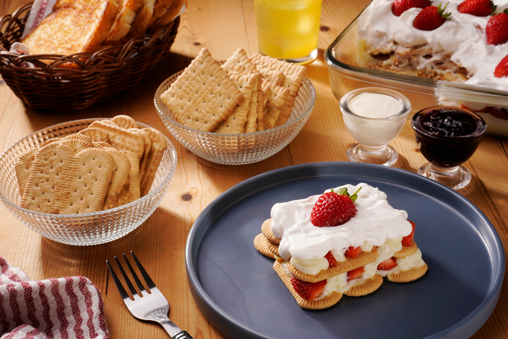 Mesa composta por biscoitos maisena, acompanhados de copo de leite, geleia, pãos tostados e sobremesa de morango, dispostos sobre mesa de madeira.