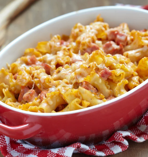 Travessa vermelha com macarrão espiral gratinado ao forno, coberto com bacon e mussarela derretida, servida sobre mesa de madeira.