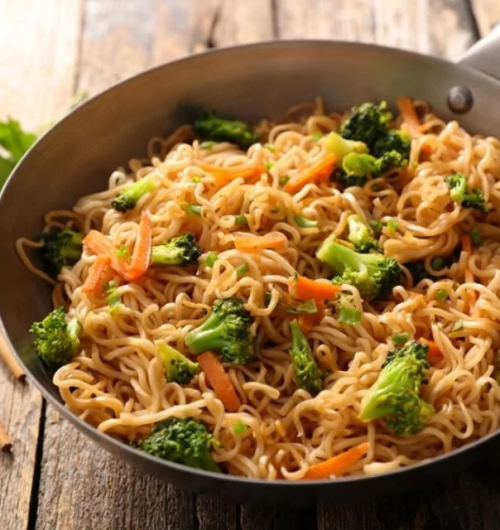 Tigela com macarrão instantâneo servido com cenoura, brócolis e pimentão, sobre mesa de madeira.