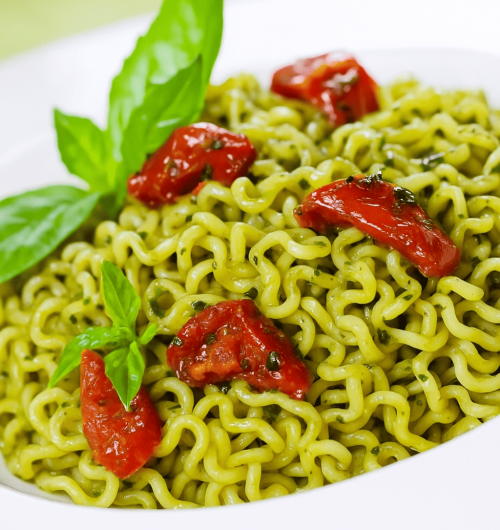 Tigela branca com macarrão instantâneo ao molho pesto, servido com pedaços de tomate e folhas de manjericão, sobre fundo desfocado.