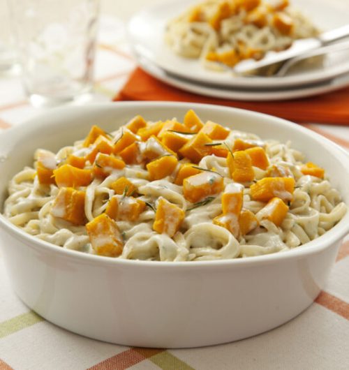 Prato com macarrão tipo ninho servido com molho de gorgonzola, acompanhado de cubos de abóbora assada e decorado com ervas, sobre mesa clara.