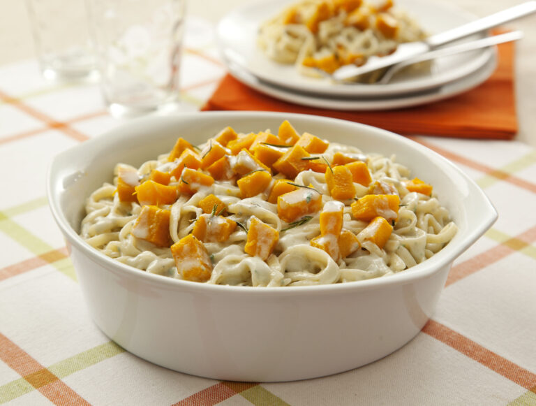 Prato com macarrão tipo ninho servido com molho de gorgonzola, acompanhado de cubos de abóbora assada e decorado com ervas, sobre mesa clara.