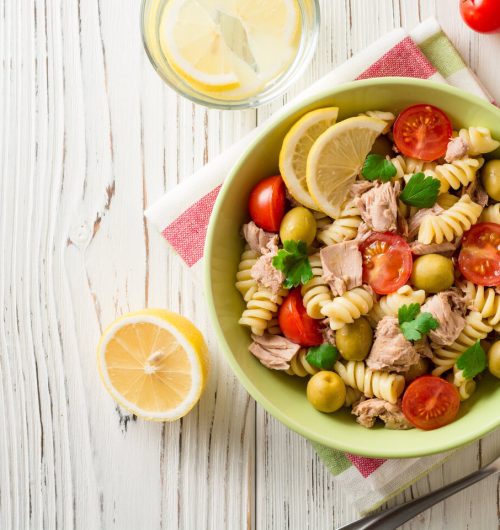 Prato com macarrão parafuso, servido com pedaços de atum, tomate, azeitona e ervas frescas, sobre mesa de madeira.