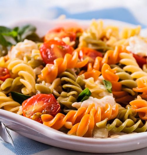 Prato de macarrão parafuso sabor vegetais servido com folhas de manjericão, frango e tomates, em prato branco sobre fundo desfocado.