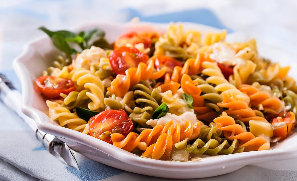 Prato de macarrão parafuso sabor vegetais servido com folhas de manjericão, frango e tomates, em prato branco sobre fundo desfocado.