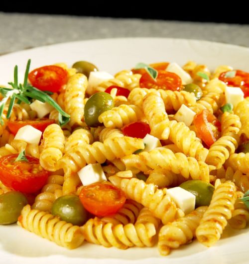 Prato de macarrão parafuso com tomates, azeitonas e queijo, servido em prato branco sobre fundo neutro.
