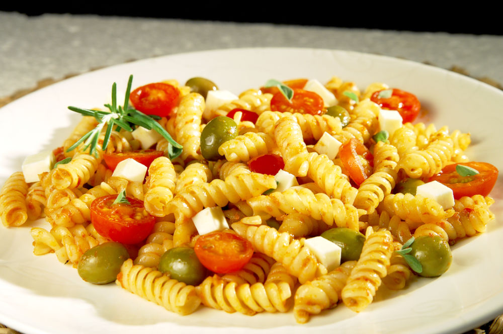 Prato de macarrão parafuso com tomates, azeitonas e queijo, servido em prato branco sobre fundo neutro.