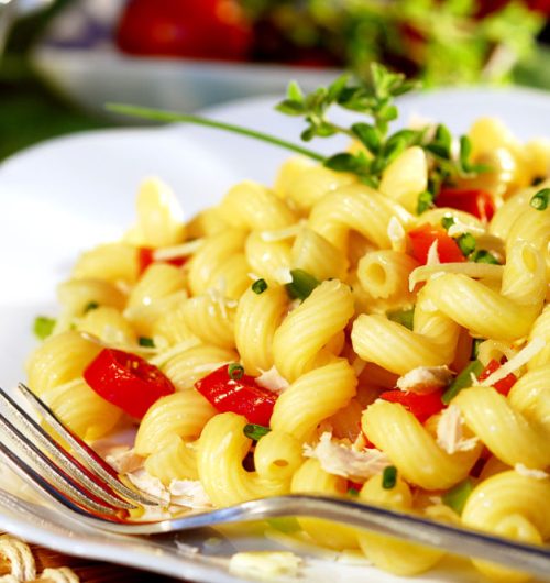 Prato de macarrão espiral com frango, tomate e pimentão verde, coberto com queijo e cebolinha, servido em prato branco; fundo desfocado.