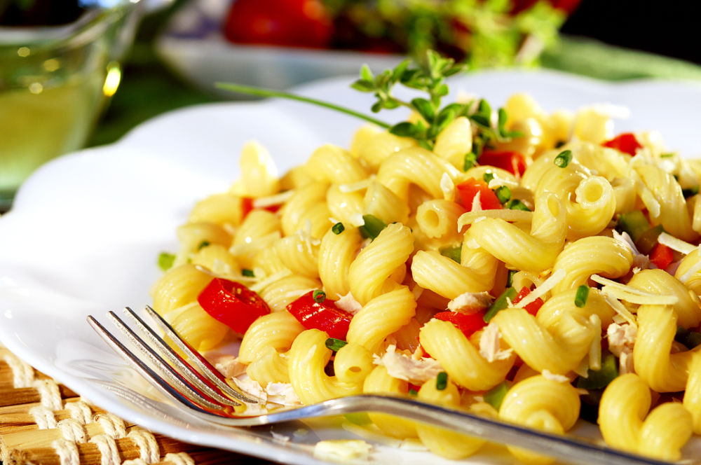 Prato de macarrão espiral com frango, tomate e pimentão verde, coberto com queijo e cebolinha, servido em prato branco; fundo desfocado.