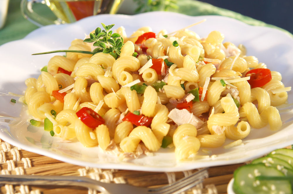 Prato de macarrão espiral com frango, tomate e pimentão verde, coberto com queijo e cebolinha, servido em prato branco; fundo desfocado.