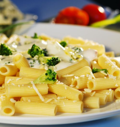 Prato de macarrão rigatoni, coberto com molho branco, brócolis e queijo ralado, disposto em um prato branco e fundo desfocado.