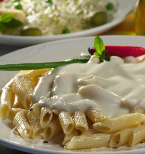 Prato de penne coberto com molho branco, pimenta e cebolinha, disposto sobre em prato branco sobre a mesa.