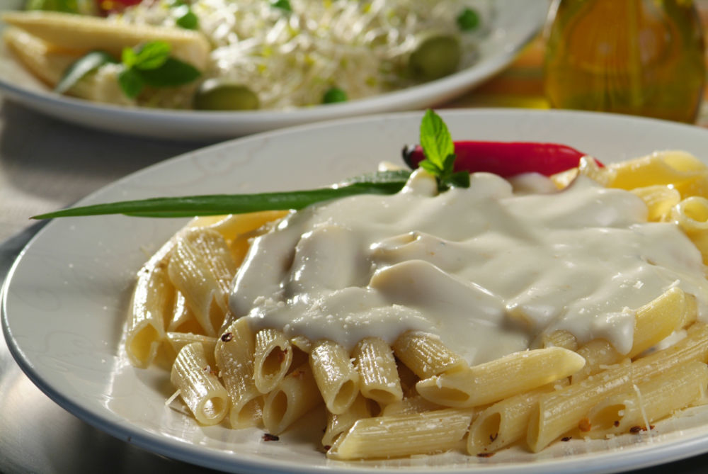 Prato de penne coberto com molho branco, pimenta e cebolinha, disposto sobre em prato branco sobre a mesa.