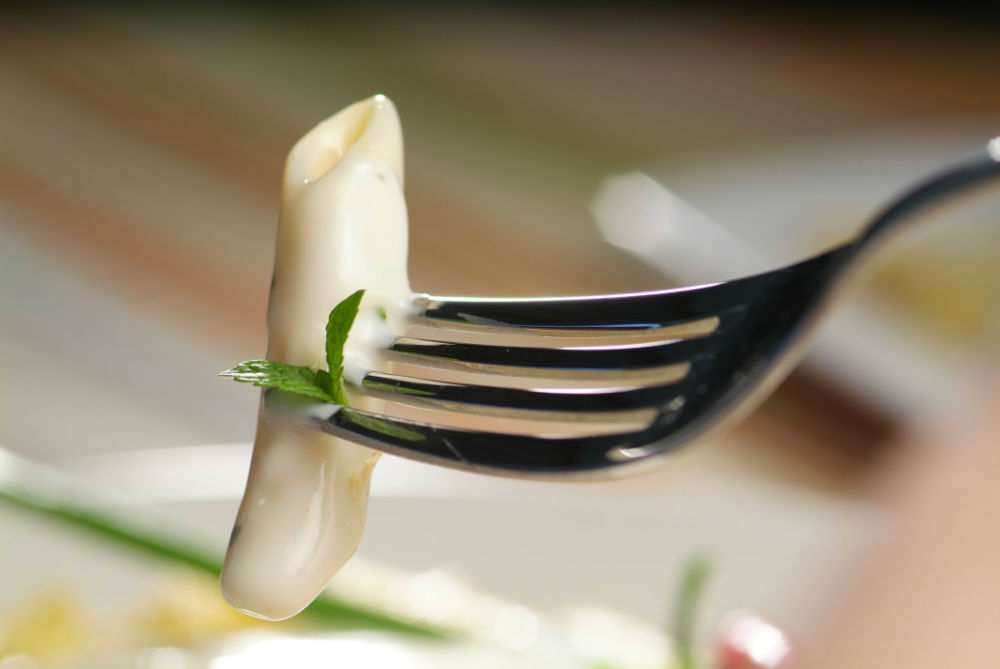 Garfo segurando macarrão penne, coberto de molho branco e uma folha de manjericão, sobre fundo desfocado.