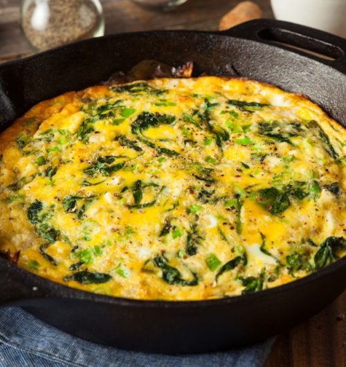 Prato de omelete com espinafre e queijo ralado, servido em frigideira sobre mesa de madeira.