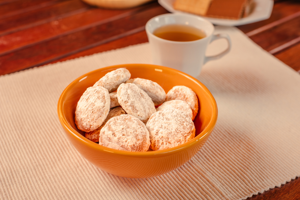 Pote amarelo com pedaços de pão de mel cobertos por açúcar, acompanhados de xícara de chá, sobre fundo iluminado