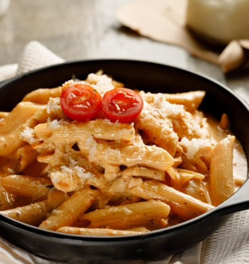 Prato fundo preto com macarrão penne ao molho cremoso, servido com queijo ralado e tomates, sobre mesa de madeira clara.
