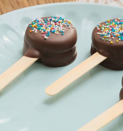 Pirulitos de biscoitos recheados cobertos de chocolate e confeitos, dispostos sobre mesa de madeira.