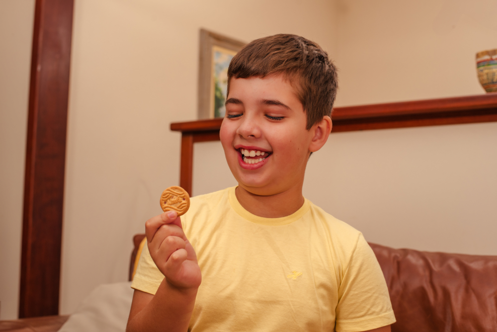 Criança sorridente segura um biscoito recheado com as mãos, em ambiente interno iluminado.