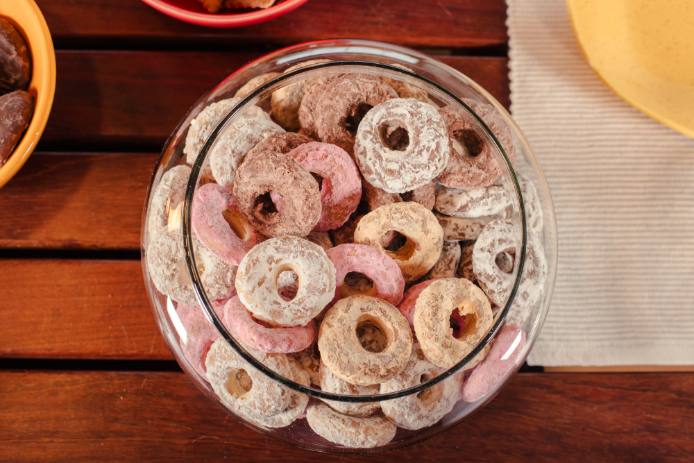 Pote com biscoitos rosca de calda sabores variados, como cappuccino, morango e pão de mel, servidos em mesa de madeira.