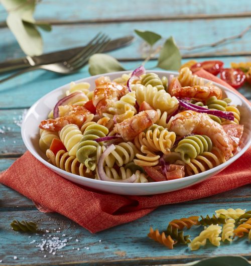 Prato branco com salada de massa tricolore, servida com camarões, tomates e cebolas roxas, sobre mesa de madeira.