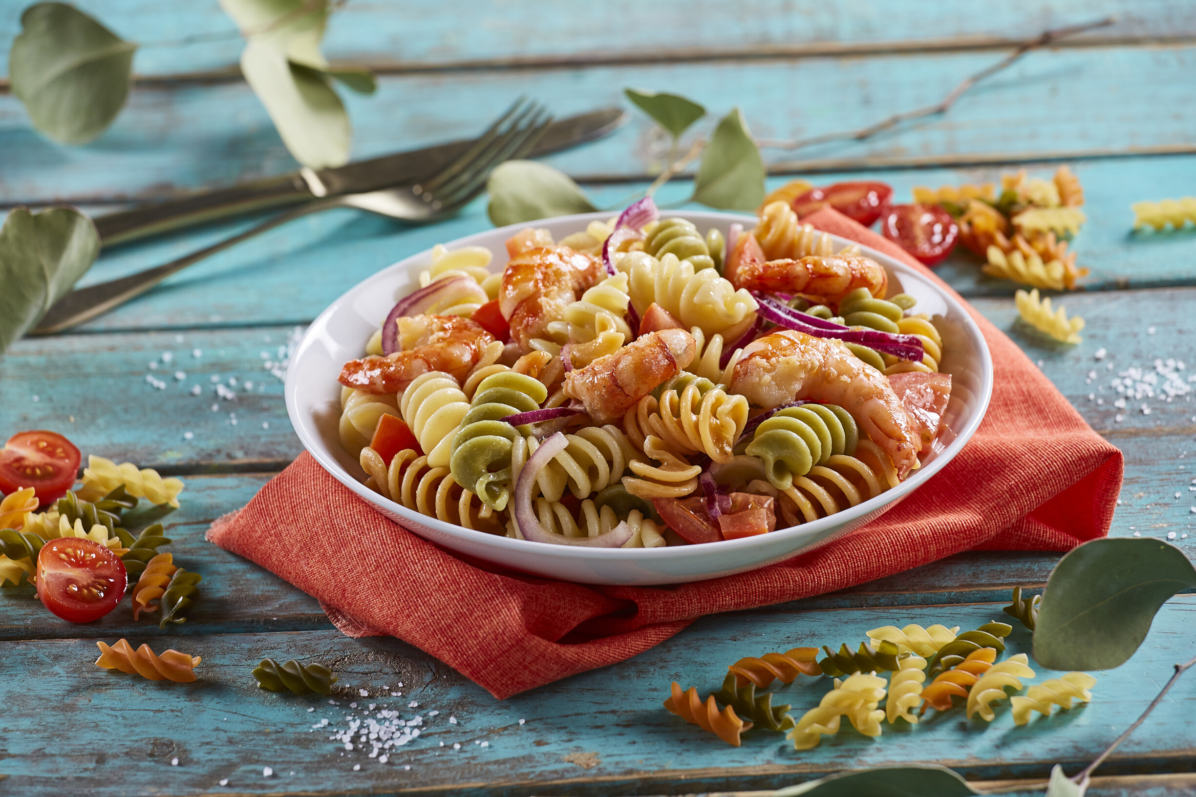 Prato branco com salada de massa tricolore, servida com camarões, tomates e cebolas roxas, sobre mesa de madeira.