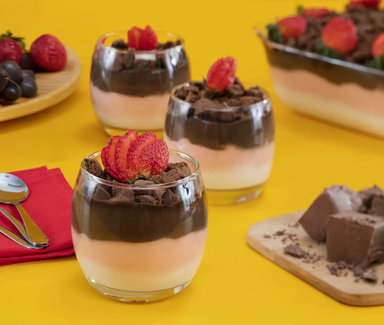 Taça de vidro transparente com sobremesa cremosa de rosquinha, decorada com morangos e calda de chocolate, sobre fundo amarelo.